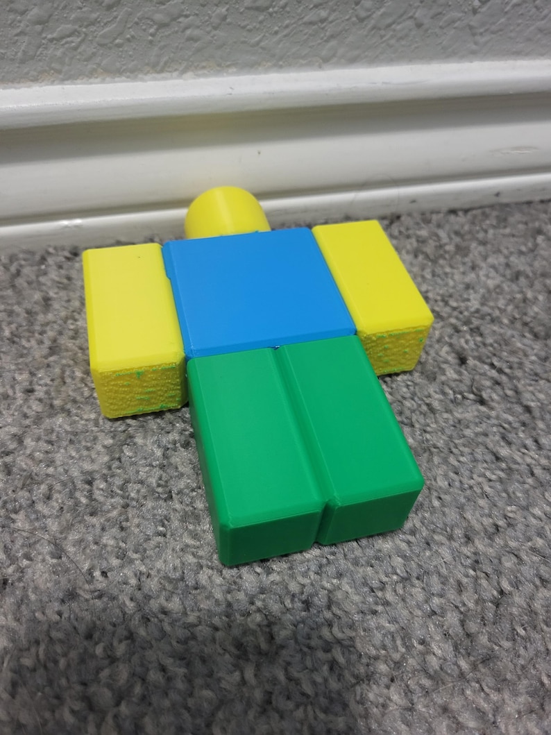 May include: A colorful, blocky figure with a yellow head and arms, a blue torso, and green legs. The figure is a popular character from a video game. The figure is resting on a gray carpet.