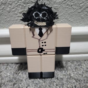 May include: A beige and black painted wooden figurine of a person wearing a trench coat, tie, and black shoes. The figurine has a black painted head with a white face and black eyes.