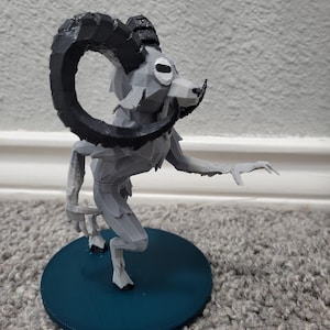 Op de afbeelding: Een grijze, 3D-geprinte figuur van een mythisch wezen met een rammenkop en grote, gebogen zwarte hoorns. De figuur staat op een teal-kleurige, cirkelvormige basis, met een uitgestrekte arm.