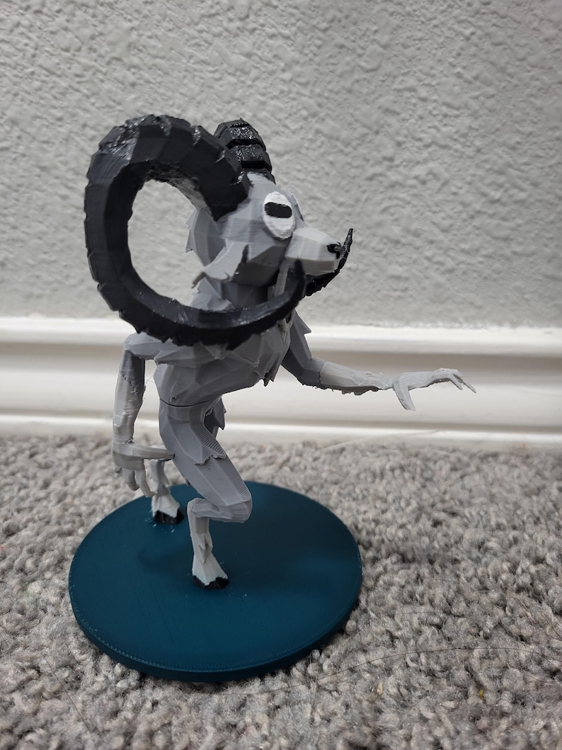 Op de afbeelding: Een grijze, 3D-geprinte figuur van een mythisch wezen met een rammenkop en grote, gebogen zwarte hoorns. De figuur staat op een teal-kleurige, cirkelvormige basis, met een uitgestrekte arm.