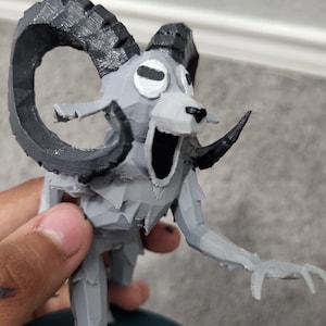 Op de afbeelding: Een grijs en zwart 3D-geprint beeldje van een ramachtig wezen met grote gebogen hoorns, een open mond en uitgestrekte armen. Het beeldje heeft een gestileerd, geometrisch ontwerp en is gemonteerd op een donkerblauwe basis.
