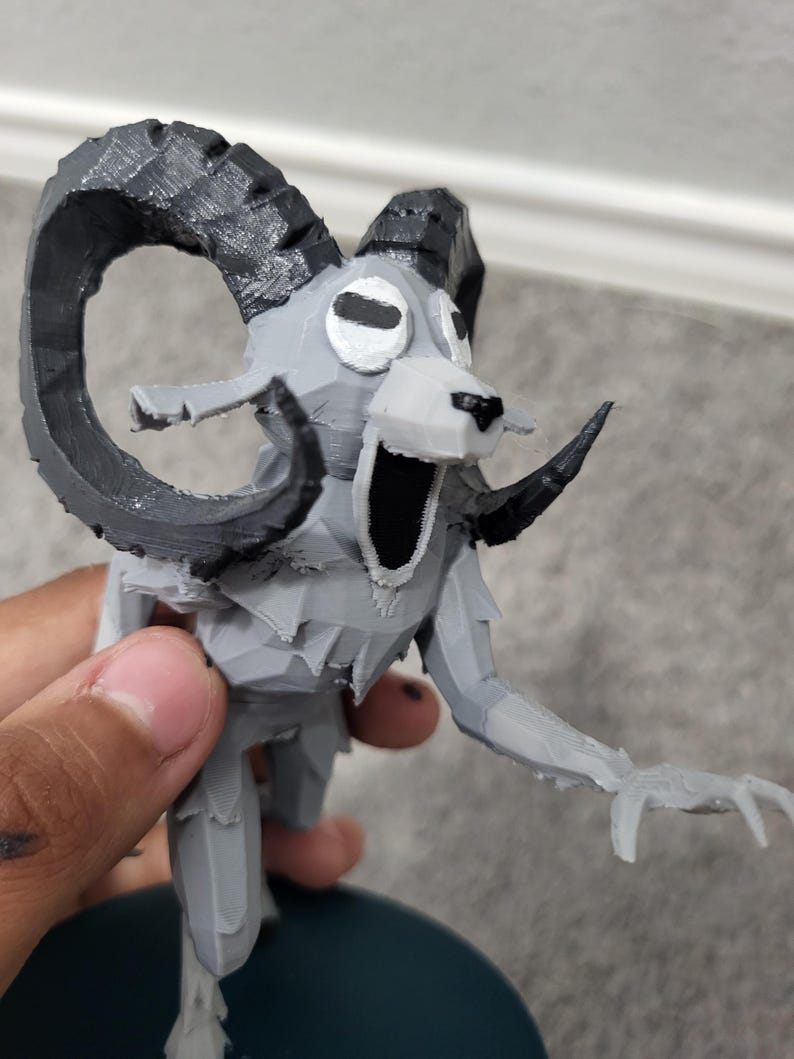 Op de afbeelding: Een grijs en zwart 3D-geprint beeldje van een ramachtig wezen met grote gebogen hoorns, een open mond en uitgestrekte armen. Het beeldje heeft een gestileerd, geometrisch ontwerp en is gemonteerd op een donkerblauwe basis.