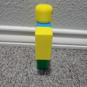 May include: A yellow, blue, and green figurine resembling a popular video game character. The figure has a yellow cylindrical head, a blue midsection, a yellow rectangular torso, and a green base. The figure is standing on a gray carpet.