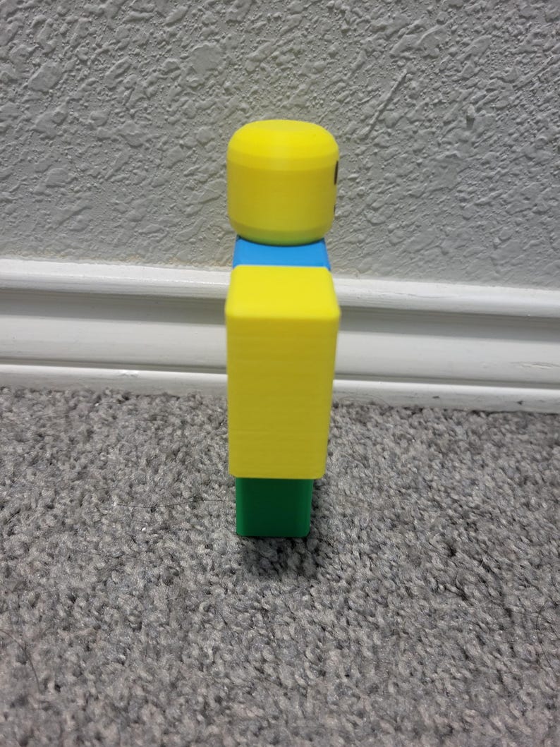 May include: A yellow, blue, and green figurine resembling a popular video game character. The figure has a yellow cylindrical head, a blue midsection, a yellow rectangular torso, and a green base. The figure is standing on a gray carpet.