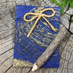 May include: A small blue notebook with a gold marbled design and a twine bow. A wooden pencil is lying next to the notebook on a wooden surface.