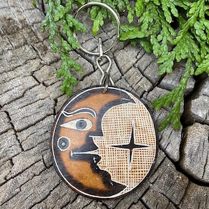 May include: A wooden keychain shaped like a crescent moon with a stylized face and a star design. The keychain is attached to a silver key ring.
