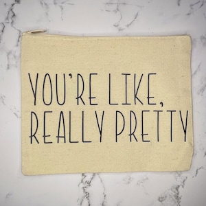 May include: A beige canvas zippered pouch with the text "YOU'RE LIKE, REALLY PRETTY" printed in black. The pouch has a gold-colored zipper and is rectangular in shape.
