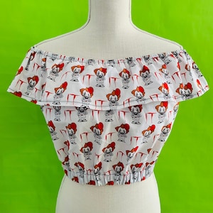 May include: White off-the-shoulder crop top with a repeating pattern of Pennywise the clown from the movie IT. The top has a ruffled neckline and a fitted bodice.