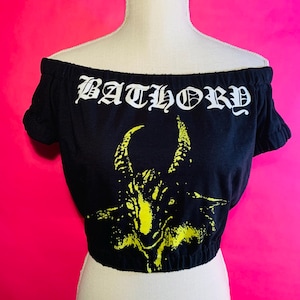 Op de afbeelding: Zwarte off-the-shoulder crop top met het woord "BATHORY" in witte gotische letters. Een gele afbeelding van een gehoornde figuur is gecentreerd onder de tekst. De top wordt getoond op een mannequin tegen een felroze achtergrond.