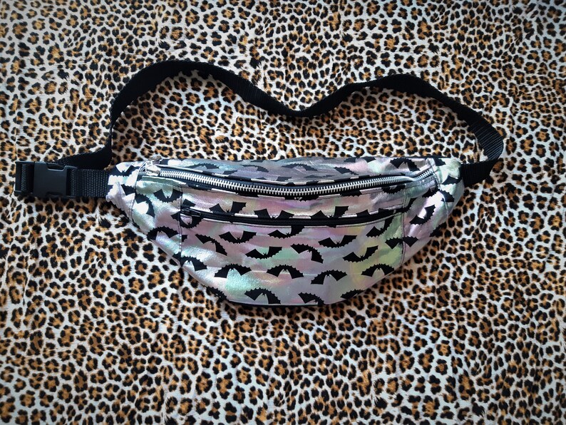 May include: A silver iridescent fanny pack with a black adjustable strap and a black bat pattern. The fanny pack is on a leopard print background.