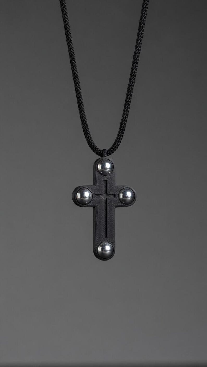 May include: A black cross pendant necklace with four silver-colored accents. The cross is suspended from a black cord. The background is a solid gray.