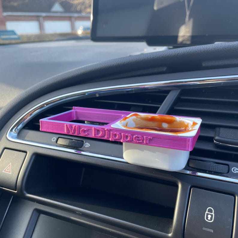 Op de afbeelding: Een roze 3D-geprinte auto-luchtroosterhouder met de woorden "Mc Dipper" erop gedrukt. Een witte dipsauscontainer met rode saus erin zit in de houder.
