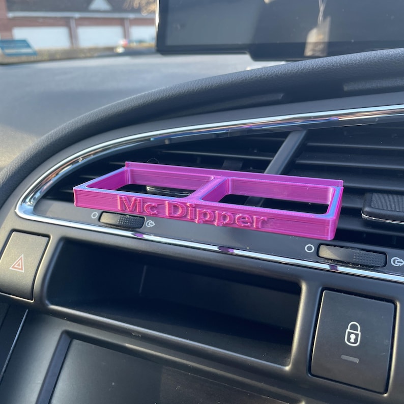 Op de afbeelding: Een roze plastic telefoonhouder voor de ventilatieopeningen in de auto met de tekst "Mc Dipper" erop gedrukt.