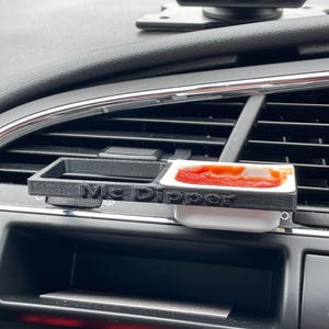 May include: A black 3D printed car air vent mount with the words "Mc Dipper" printed on it. A small white condiment dish with red sauce is in the mount.