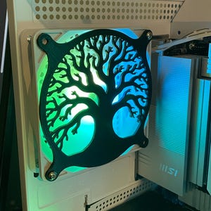 May include: A black tree of life design covers a computer fan, mounted on a white computer case. The fan glows with green and blue LED lights. The computer case has a GeForce RTX sticker.