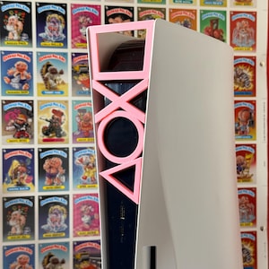 Può includere: Una console di gioco bianca e nera con simboli rosa del controller sul lato. La console è posizionata di fronte a un muro coperto di figurine vintage di Garbage Pail Kids.