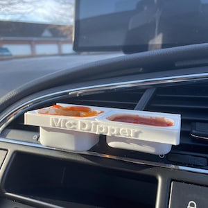 Op de afbeelding: Een witte plastic McDipper-container met drie compartimenten, waarvan er twee gevuld zijn met rode saus. De container zit in de ventilatieopening van een auto.