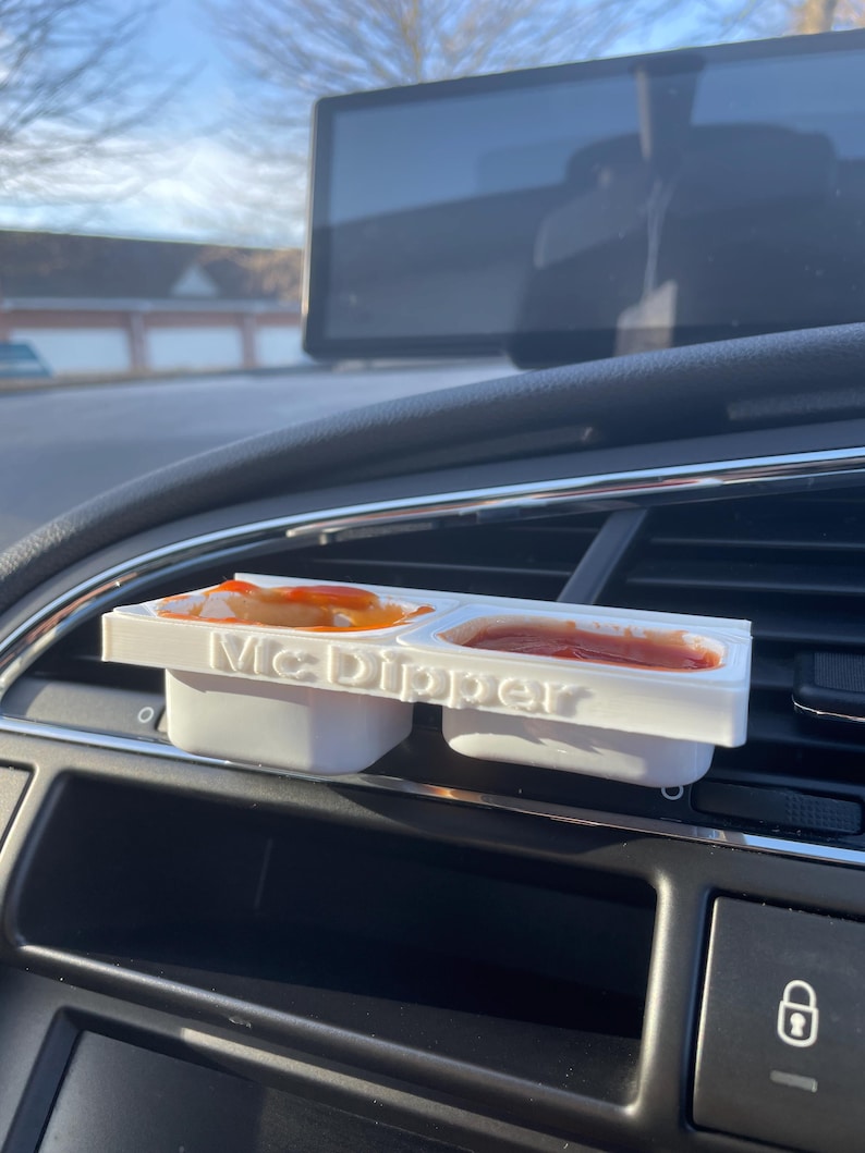 Op de afbeelding: Een witte plastic McDipper-container met drie compartimenten, waarvan er twee gevuld zijn met rode saus. De container zit in de ventilatieopening van een auto.