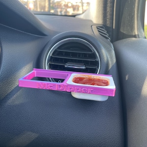 Op de afbeelding: Een roze 3D-geprinte auto-luchtroosterhouder met de woorden "Mc Dipper" erop gedrukt. Een kleine witte plastic container met rode saus zit in de houder.