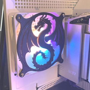 Puede incluir: Una cubierta de ventilador negra con forma de dragón está montada en una caja de ordenador. El ventilador brilla con un arcoíris de colores. La caja del ordenador es blanca y los componentes son visibles en el interior.