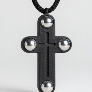 May include: A black cross pendant with four silver spheres at the ends, suspended from a black cord. The cross has a textured surface and a simple, modern design. The pendant is a religious symbol.
