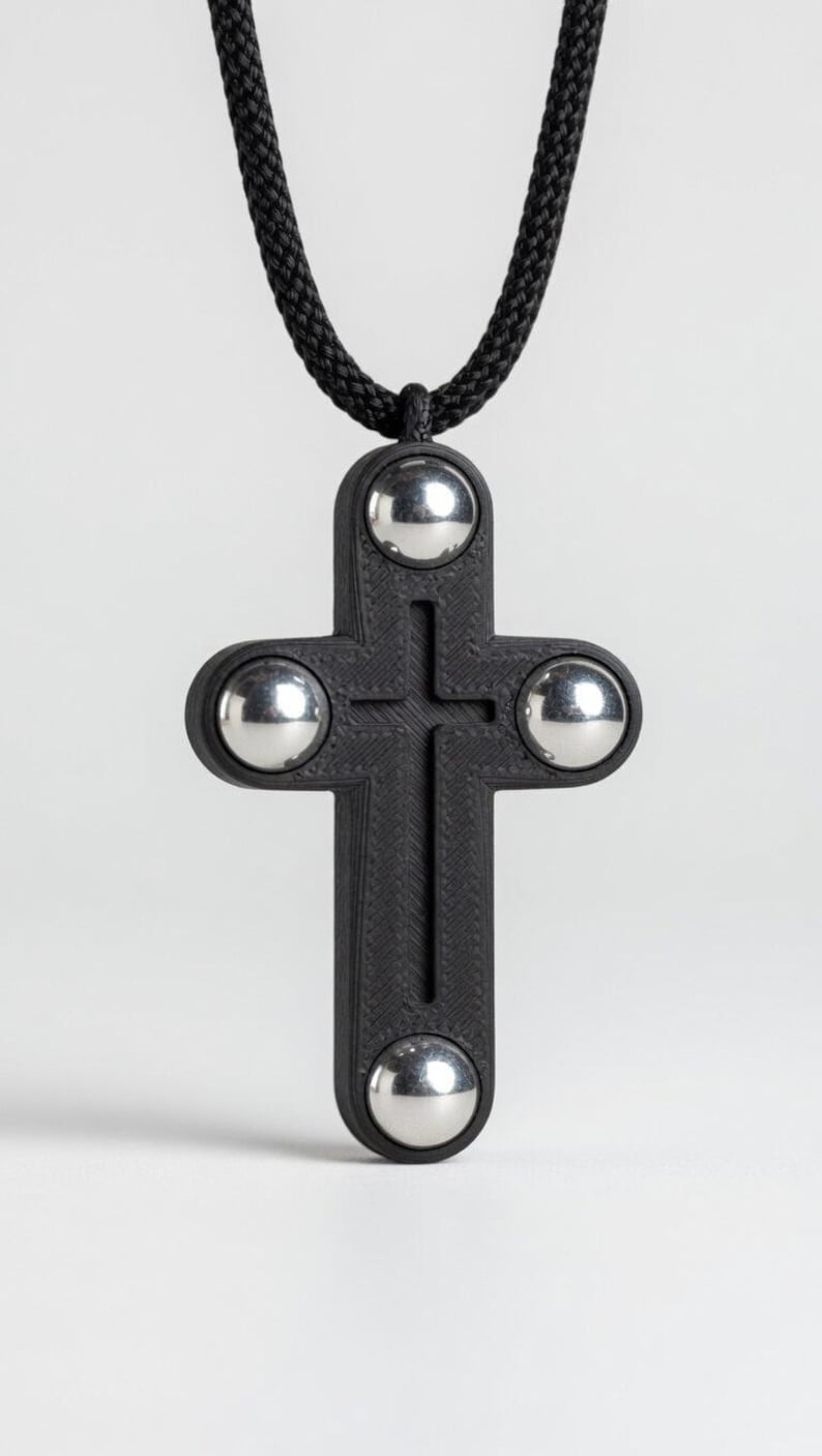 May include: A black cross pendant with four silver spheres at the ends, suspended from a black cord. The cross has a textured surface and a simple, modern design. The pendant is a religious symbol.