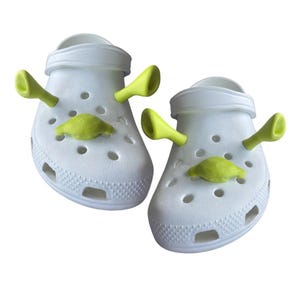 Könnte beinhalten: Ein Paar weiße Clogs mit limettengrünen dekorativen Shrek-Anhängern. Die Anhänger zeigen die Ohren und das Gesicht der Figur. Die Clogs haben mehrere Löcher und eine strukturierte Oberfläche.