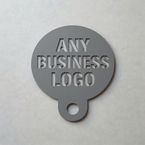 May include: A grey, circular stencil with the words "ANY BUSINESS LOGO" cut out. The stencil has a small tab with a hole at the bottom. The background is a light grey colour.