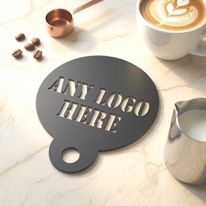 May include: A circular black coffee stencil featuring the laser-cut text ANY LOGO HERE. The stencil includes a small handle with a circular hole at the bottom, designed for creating decorative patterns on the foam of coffee beverages.