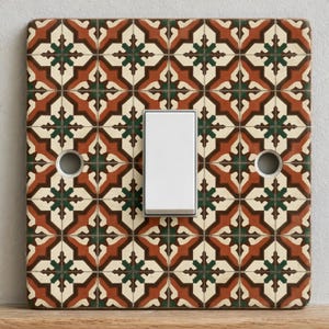 May include: A square light switch cover plate featuring a repeating geometric pattern in shades of terracotta, dark green, and cream. The plate has a centred white rectangular rocker switch and two circular screw holes on the left and right sides.