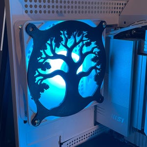 Op de afbeelding: Een computerkastventilator met een zwart levensboomontwerp. De ventilator wordt verlicht door een blauw LED-licht. De computerkast is wit.