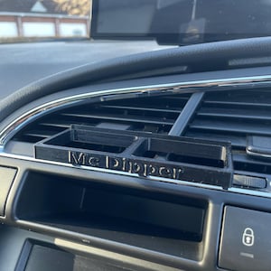 Op de afbeelding: Een zwarte 3D-geprinte autotelefoonhouder met de woorden "Mc Dipper" erop gedrukt. De houder is bevestigd aan de ventilatieopening van een auto.