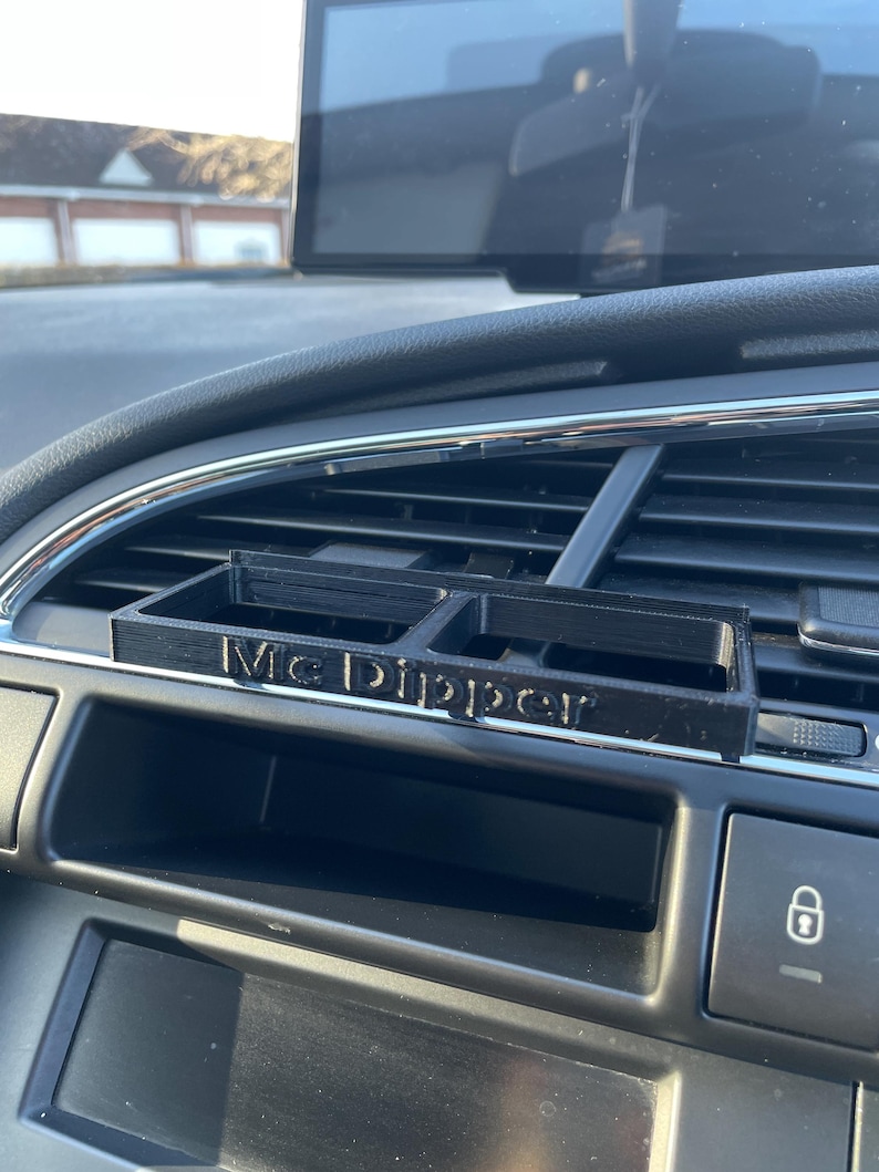 Op de afbeelding: Een zwarte 3D-geprinte autotelefoonhouder met de woorden "Mc Dipper" erop gedrukt. De houder is bevestigd aan de ventilatieopening van een auto.