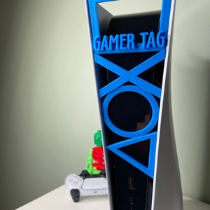 May include: A white PlayStation 5 console with a blue 3D printed gamer tag holder. The holder features the PlayStation symbols and the text "GAMER TAG".