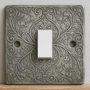 May include: A square light switch cover plate featuring a raised, weathered grey metal finish with an intricate embossed floral and vine pattern. The plate has two circular screw holes on the sides and a centred, rectangular white rocker switch.