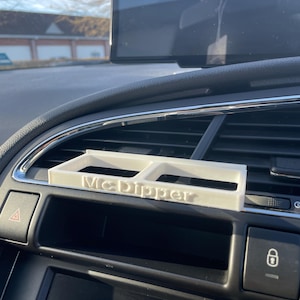 Op de afbeelding: Een witte plastic autotelefoonhouder met de tekst "McDipper" erop gedrukt. De houder is ge&iuml;nstalleerd in een autoventilatieopening.
