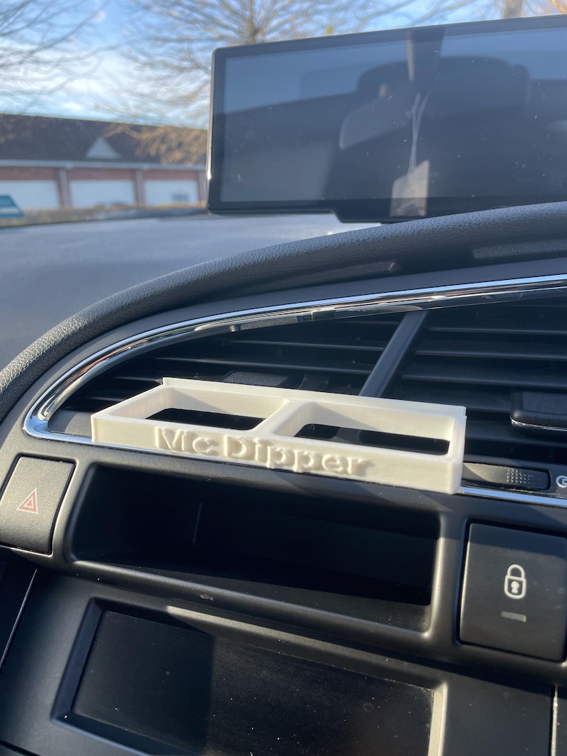 Op de afbeelding: Een witte plastic autotelefoonhouder met de tekst "McDipper" erop gedrukt. De houder is ge&iuml;nstalleerd in een autoventilatieopening.
