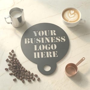 May include: A circular dark grey stencil featuring the cut-out text "YOUR BUSINESS LOGO HERE" with a circular handle at the bottom. The stencil is positioned alongside a silver milk pitcher, a white coffee cup with latte art, and coffee beans.
