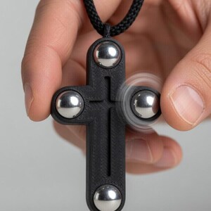 May include: A black cross-shaped pendant with four silver ball bearings. The pendant is suspended from a black cord. One of the ball bearings is spinning, creating a blur effect. The pendant is held in a person's hand.