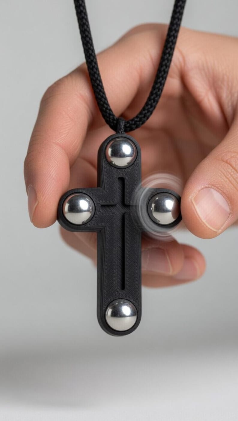 May include: A black cross-shaped pendant with four silver ball bearings. The pendant is suspended from a black cord. One of the ball bearings is spinning, creating a blur effect. The pendant is held in a person's hand.