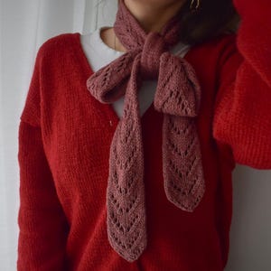 Könnte beinhalten: Ein altrosa gestrickter Schal mit dekorativem Lochmuster liegt über einer roten Strickjacke. Der Schal ist zu einer Schleife gebunden. Die Strickjacke hat einen V-Ausschnitt und ist an der Taille geknöpft. Ein weißes Hemd ist darunter sichtbar.