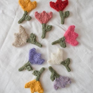Könnte beinhalten: Eine Sammlung handgefertigter gestrickter Blumenapplikationen in verschiedenen Farben, darunter Gelb, Rot, Rosa, Beige, Lila und Weiß. Jede Blume hat einen grünen Stiel und Blätter. Die Applikationen sind auf einer weißen Oberfläche angeordnet.