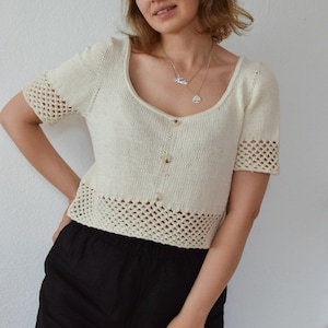 Puede incluir: Un top blanco de crochet con mangas cortas y cierre de botón. El top tiene un ribete de crochet alrededor del escote, las mangas y el dobladillo. La modelo lleva pantalones cortos negros.