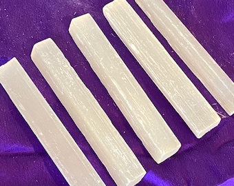 May include: Five white selenite sticks on a purple background. The sticks are smooth and have a natural, striated texture.