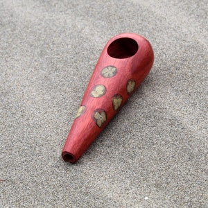 May include: A red wooden pipe with a floral design. The pipe is lying on a sandy surface.