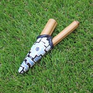 May include: A handmade wooden musical instrument, a panpipe, rests on green grass. The panpipe features a decorative, painted section with a blue and white geometric design. The two wooden pipes are light brown.