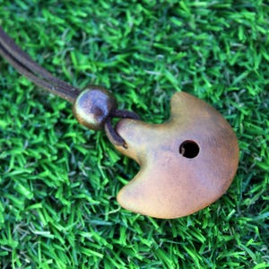 Può includere: Una collana con ciondolo in legno fatto a mano con una forma a mezzaluna. Il ciondolo è di colore marrone caldo con un piccolo foro. La collana ha un cordino marrone scuro e una perla rotonda. La collana è esposta su una superficie verde.