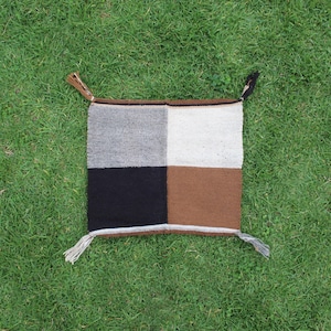 May include: A square, hand-woven pillow with a patchwork design. The pillow is divided into four squares: gray, white, black, and brown. Each corner has a tassel. The pillow is placed on a bed of green grass.