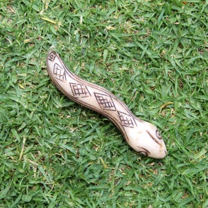 Può includere: Un serpente bianco intagliato con un motivo geometrico, sdraiato su un letto di erba verde.