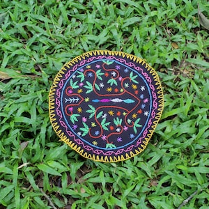 May include: A round, black fabric coaster with colourful embroidery depicting a stylised design of leaves and flowers. The coaster has a yellow, pink, and white border.
